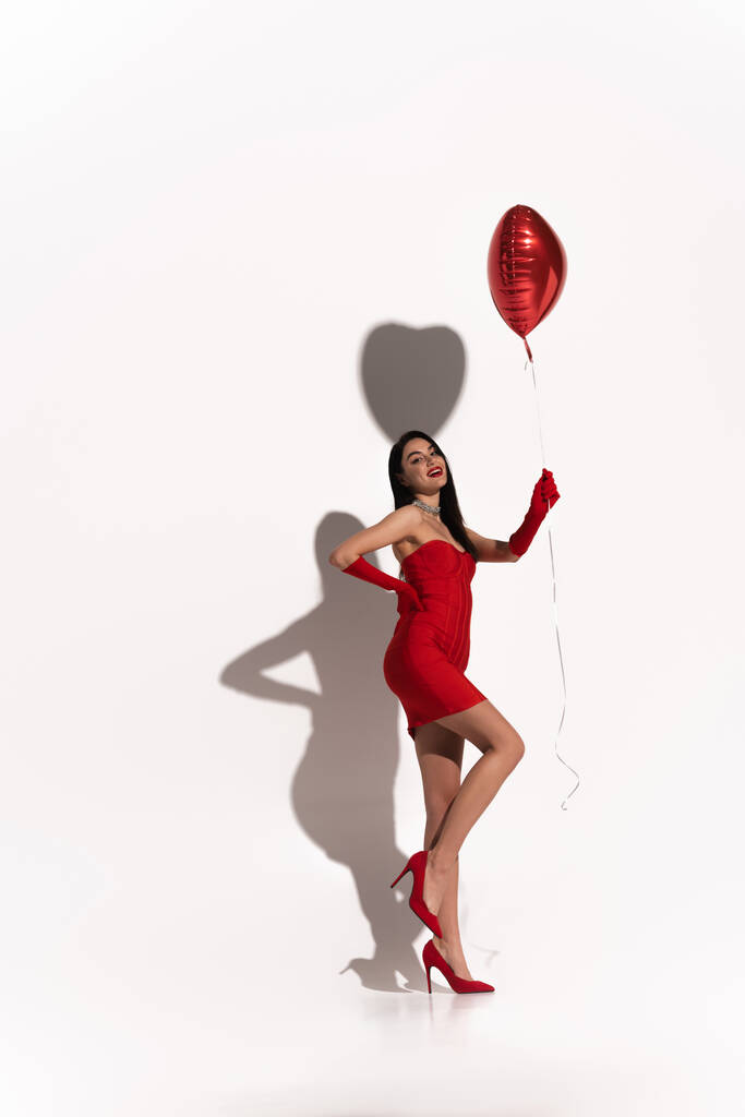 Kırmızı elbiseli ve eldivenli, beyaz arka planda kalp şeklinde balon tutan seksi bir kadın.  - Fotoğraf, Görsel