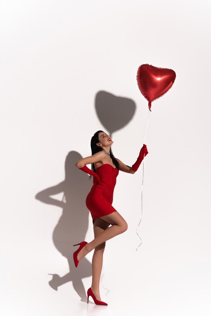 Stylish woman in red heels and dress posing and looking at heart shaped balloon on white background with shadow  - Photo, Image