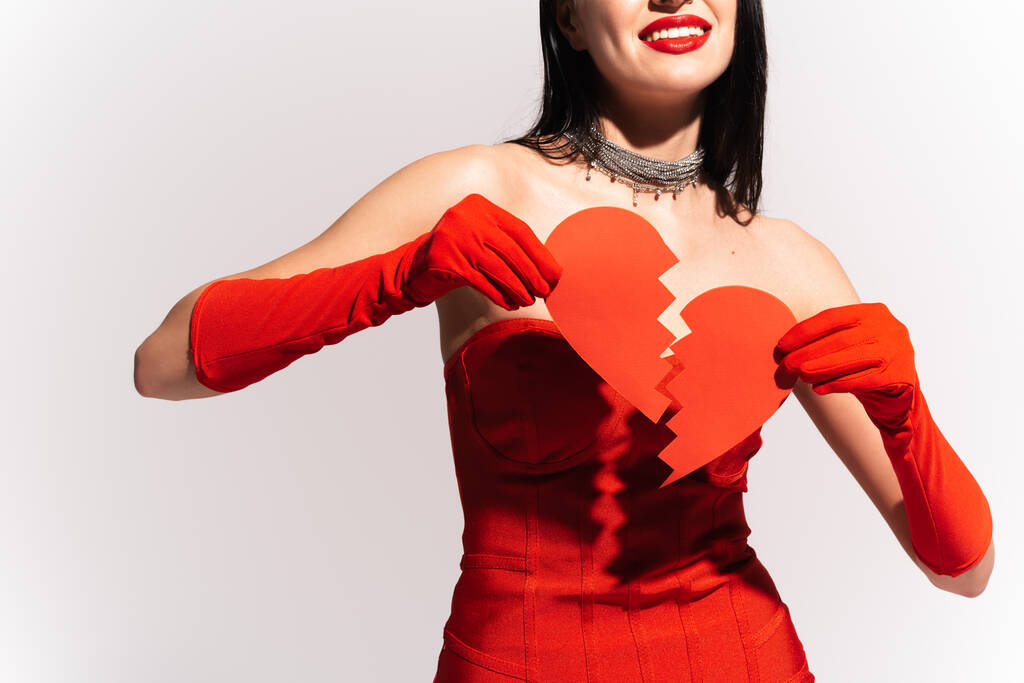 Обрізаний вид усміхненої жінки з червоними губами, що тримає папір зламаним серцем ізольовано на сірому
  - Фото, зображення