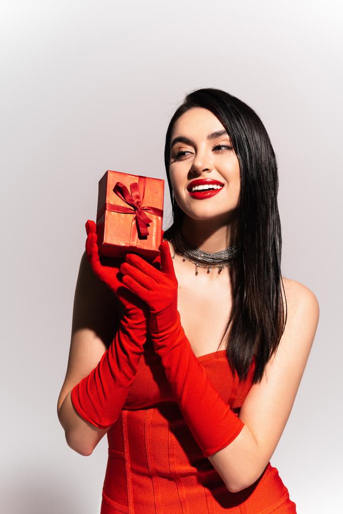 Stilvolle brünette Frau in roten Handschuhen hält Geschenkbox auf grauem Hintergrund  - Foto, Bild