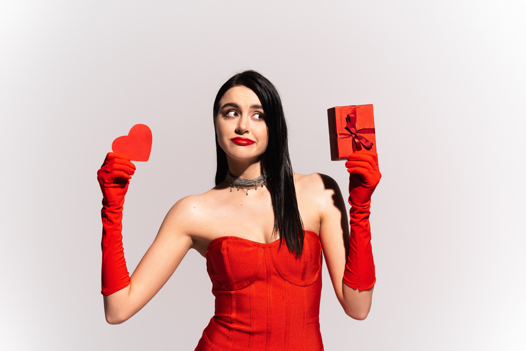 Töprengő nő piros ruhában tartó papír szív és ajándék elszigetelt szürke  - Fotó, kép