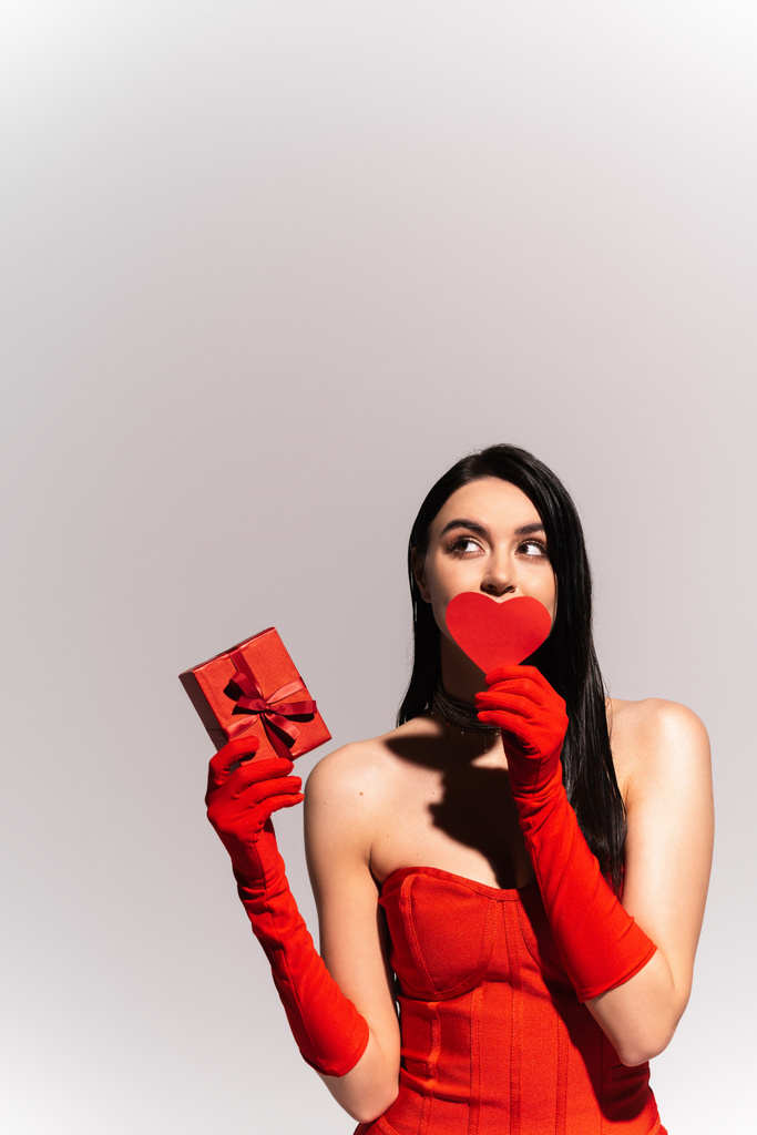 Stylowa brunetka w czerwonych rękawiczkach trzymająca papierowe serce i prezent odizolowany na szaro  - Zdjęcie, obraz