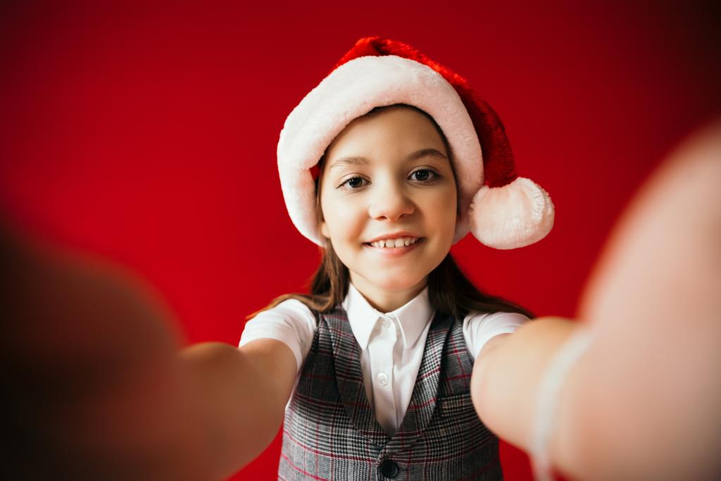joyful girl in santa hat and plaid vest smiling at camera on blurred foreground isolated on red - Photo, Image
