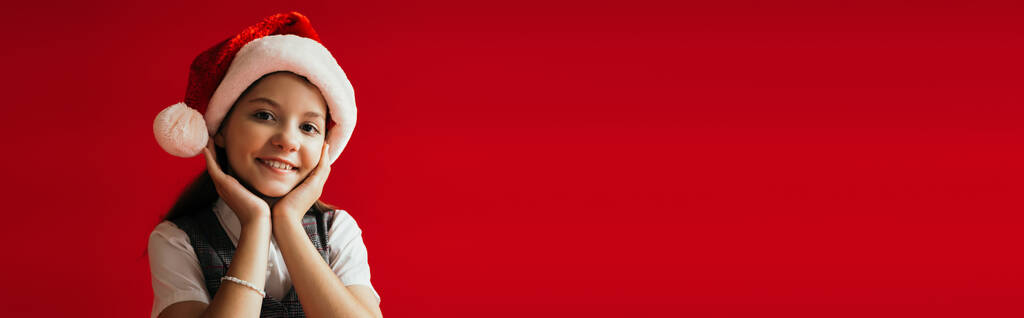 alegre chica en santa hat sonriendo a la cámara mientras sostiene las manos cerca de la cara aislado en rojo, pancarta - Foto, Imagen