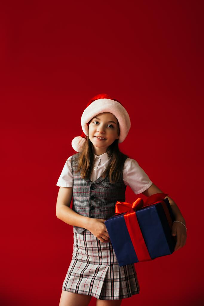 усміхнена дитина в капелюсі Санта і спідниця стоїть з різдвяним подарунком ізольовано на червоному
 - Фото, зображення