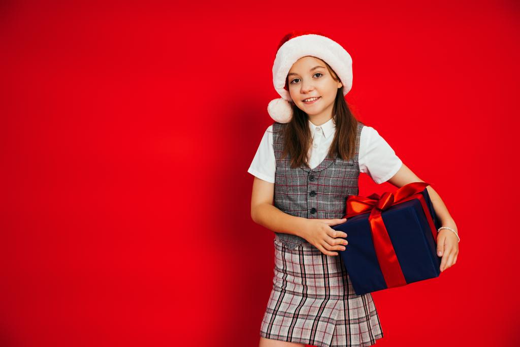 Frühchen-Mädchen in stylischer karierter Kleidung und Weihnachtsmütze mit Weihnachtsgeschenk auf rotem Grund - Foto, Bild