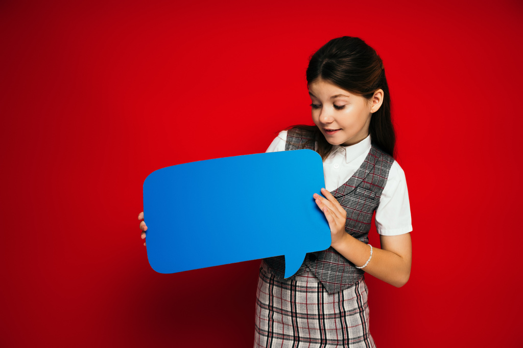 niña preadolescente sonriente en falda a cuadros mirando burbuja de habla azul aislado en rojo - Foto, imagen