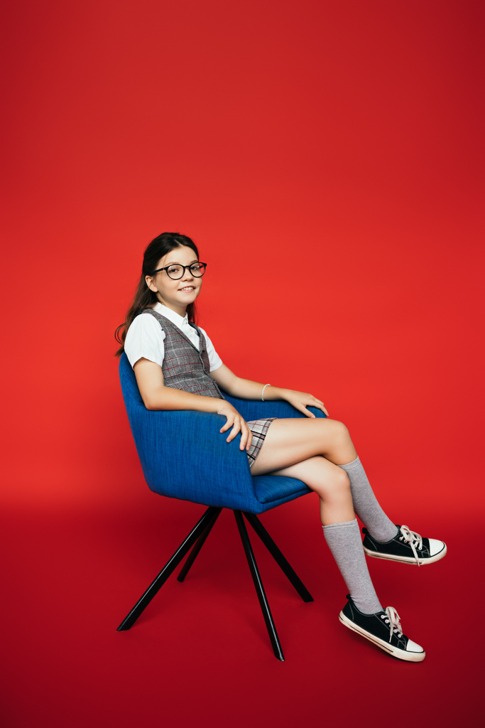 полная длина улыбающейся девушки в очках и ботинках, сидящей в синем кресле на красном фоне - Фото, изображение