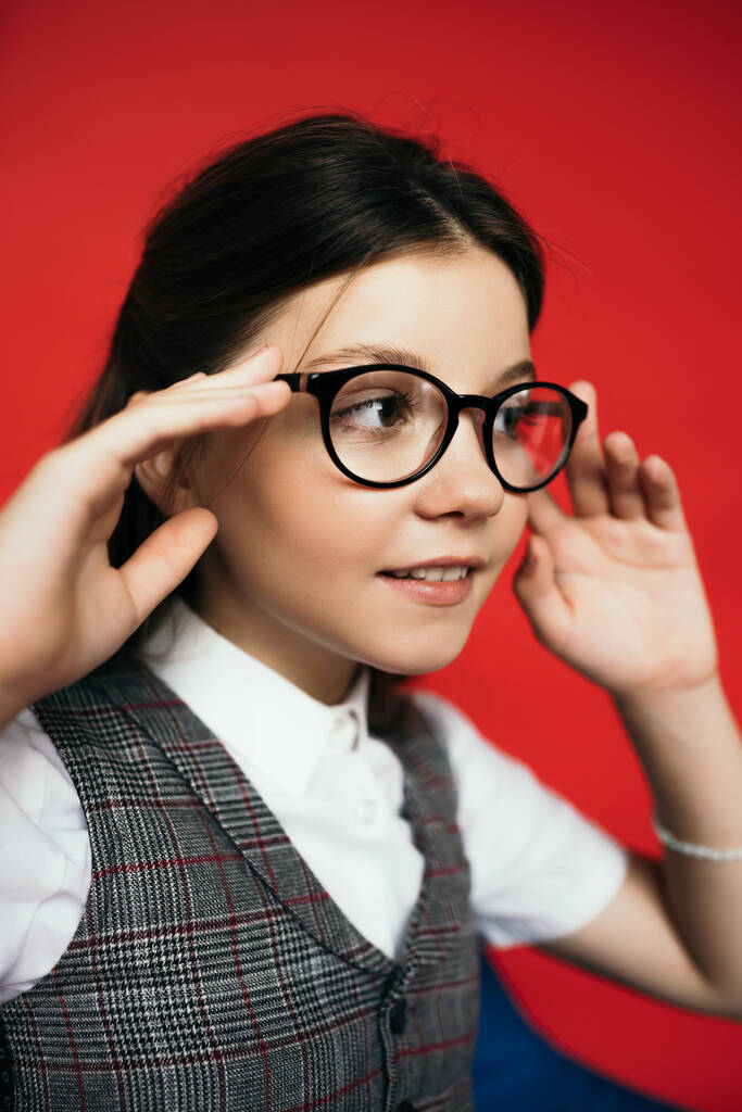 preadolescente chica ajustando gafas y sonriendo mientras mira hacia otro lado aislado en rojo - Foto, Imagen