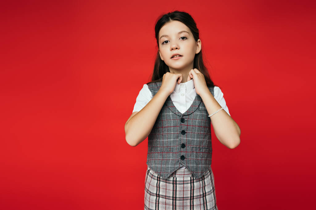 menina pré-adolescente em colete xadrez e saia ajustando colarinho de blusa branca isolada no vermelho, banner  - Foto, Imagem