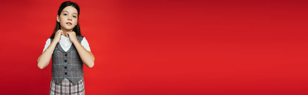 preteen ragazza bruna in gilet a scacchi regolazione del colletto della camicetta e guardando la fotocamera isolata sul rosso, banner  - Foto, immagini