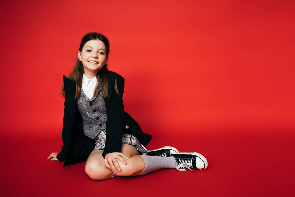 Kırmızı arka planda oturan siyah ceketli ve spor ayakkabılı mutlu bir kız. - Fotoğraf, Görsel