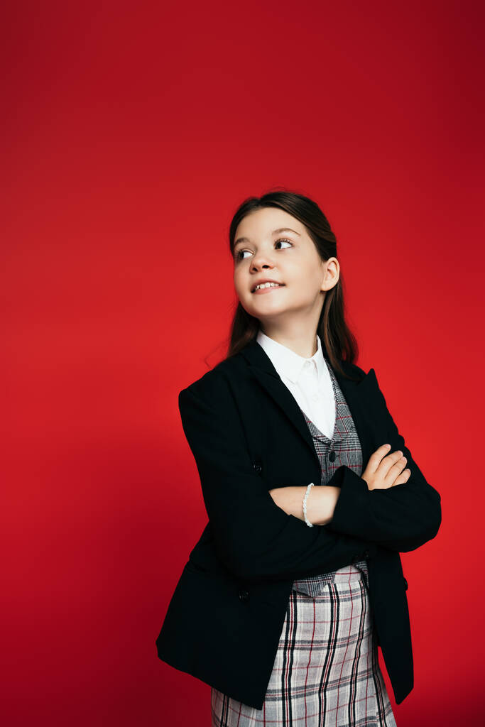 menina feliz em saia quadriculada e blazer preto de pé com braços cruzados e olhando para trás isolado no vermelho - Foto, Imagem