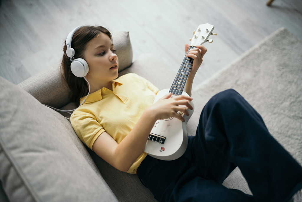 ソファに横たわって小さなハワイのギターを演奏する有線ヘッドフォンの女の子のトップビュー - 写真・画像