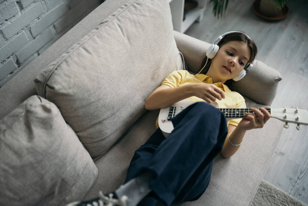 vista dall'alto della ragazza che gioca ukulele mentre si trova sul divano in cuffie cablate - Foto, immagini