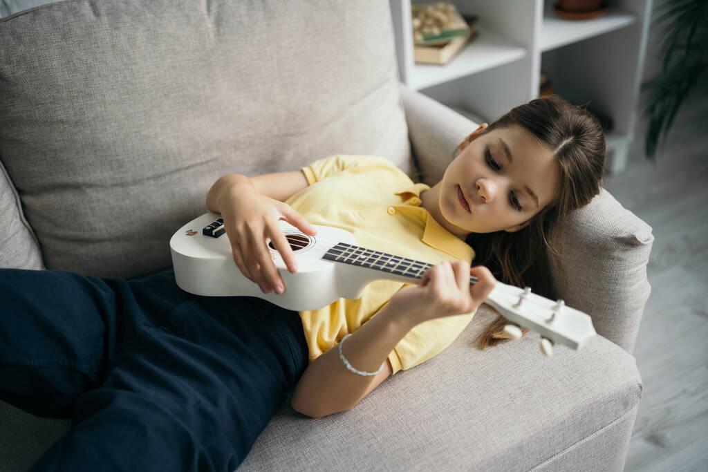 visão de alto ângulo da menina pré-adolescente deitada no sofá em casa e tocando pequena guitarra havaiana - Foto, Imagem