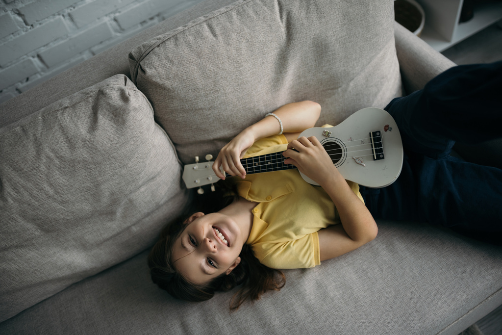 vista dall'alto di gioiosa ragazza sdraiata sul divano e suonare la piccola chitarra hawaiana - Foto, immagini