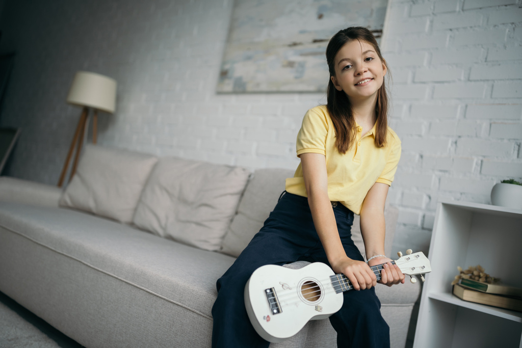 happy preteen girl holding ukulele and looking at camera near blurred couch at home - 写真・画像