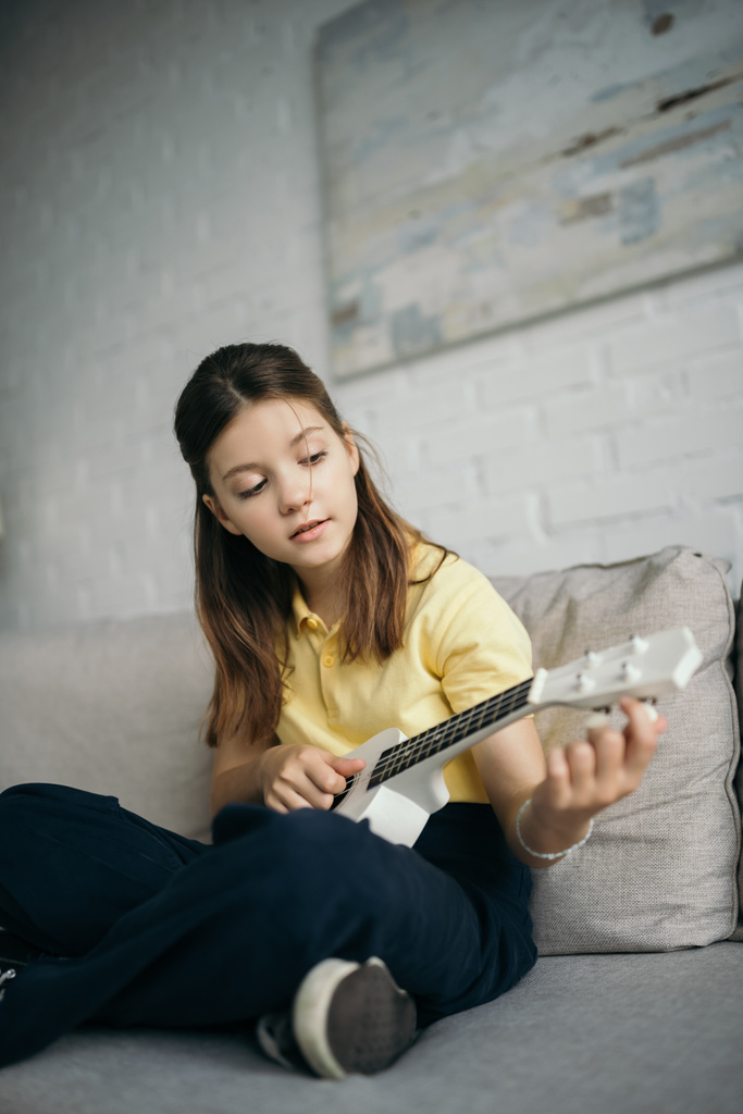 morena menina tuning pouco havaiano guitarra enquanto sentado no sofá em casa - Foto, Imagem