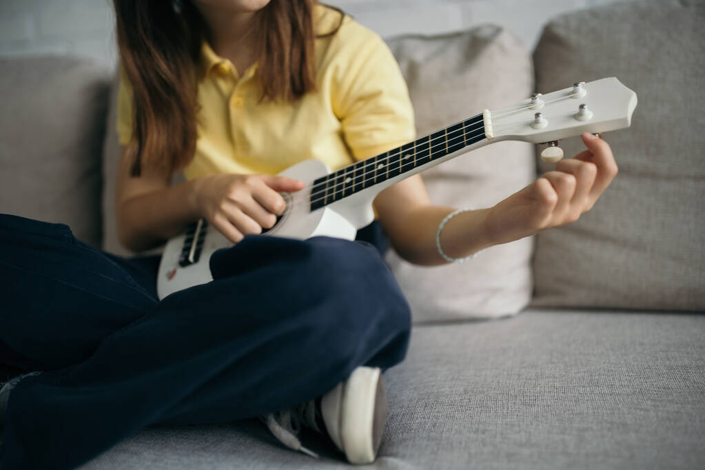 vista ritagliata di bambino offuscata tuning ukulele mentre seduto sul divano con gambe incrociate - Foto, immagini
