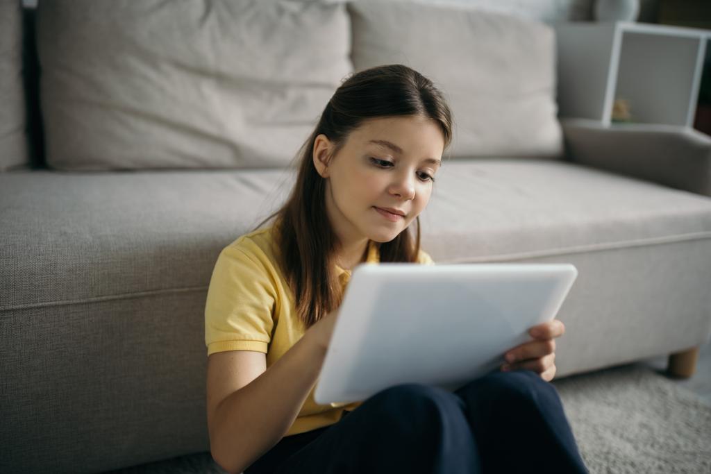 pozytywny nastolatek dziewczyna za pomocą cyfrowy tablet w pobliżu niewyraźne kanapa w salonie - Zdjęcie, obraz