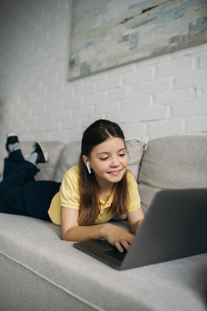 fille souriante dans un écouteur sans fil couché sur un canapé doux et tapant sur un ordinateur portable - Photo, image