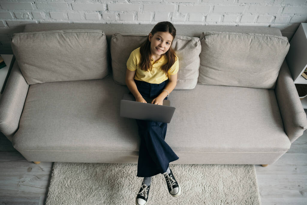 widok z góry uśmiechnięty nastolatek dziewczyna siedzi na kanapie z laptopem i patrząc w aparat - Zdjęcie, obraz