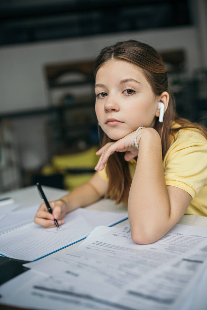 Preteen Mädchen im drahtlosen Kopfhörer hält Stift in der Hand und schaut in die Kamera, während sie Hausaufgaben macht - Foto, Bild