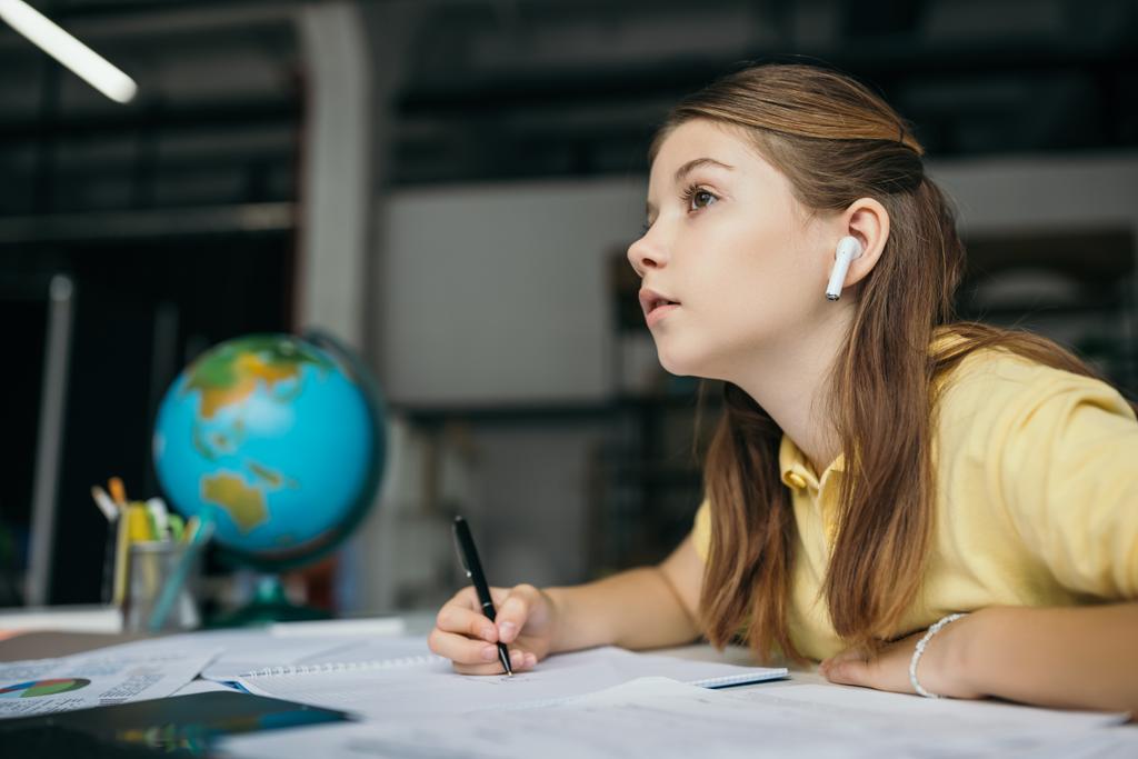 πλαϊνή άποψη του σκεπτόμενου κοριτσιού στο ασύρματο ακουστικό κρατώντας στυλό και κοιτάζοντας μακριά, ενώ κάνει την εργασία - Φωτογραφία, εικόνα
