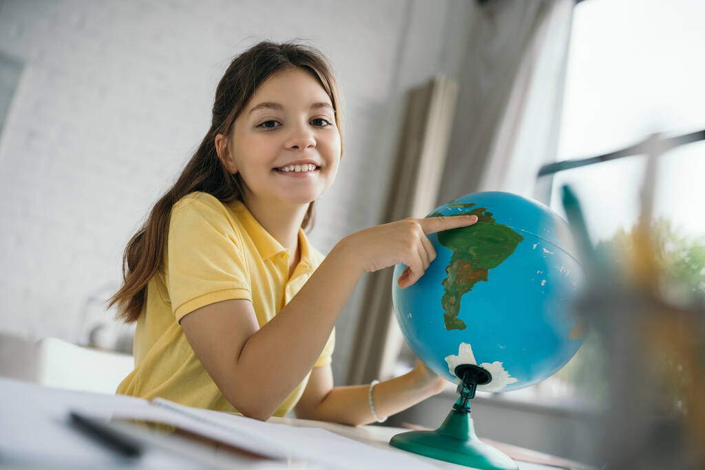 веселая девушка, указывающая на глобус и улыбающаяся в камеру во время домашнего обучения на размытом переднем плане - Фото, изображение