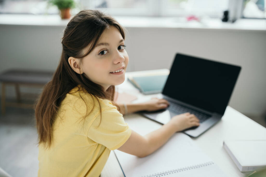 brunette meisje glimlachen op camera en typen op wazig laptop tijdens het studeren thuis - Foto, afbeelding