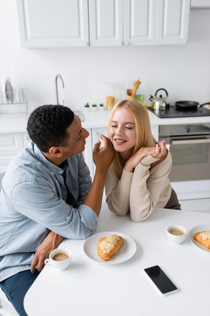 afro-amerikai férfi megható arc csinos szőke barátnő közelében kávé csésze és croissant a konyhában - Fotó, kép