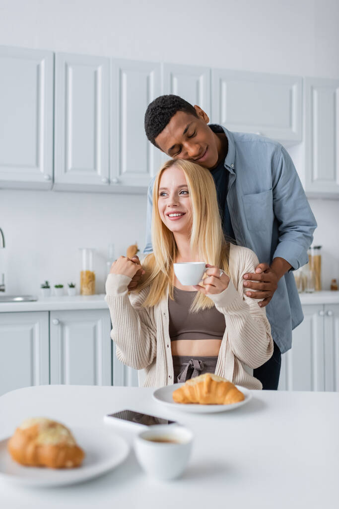 africký Američan muž se zavřenýma očima objímající blondýny přítelkyně držící kávu v blízkosti lahodné croissant - Fotografie, Obrázek