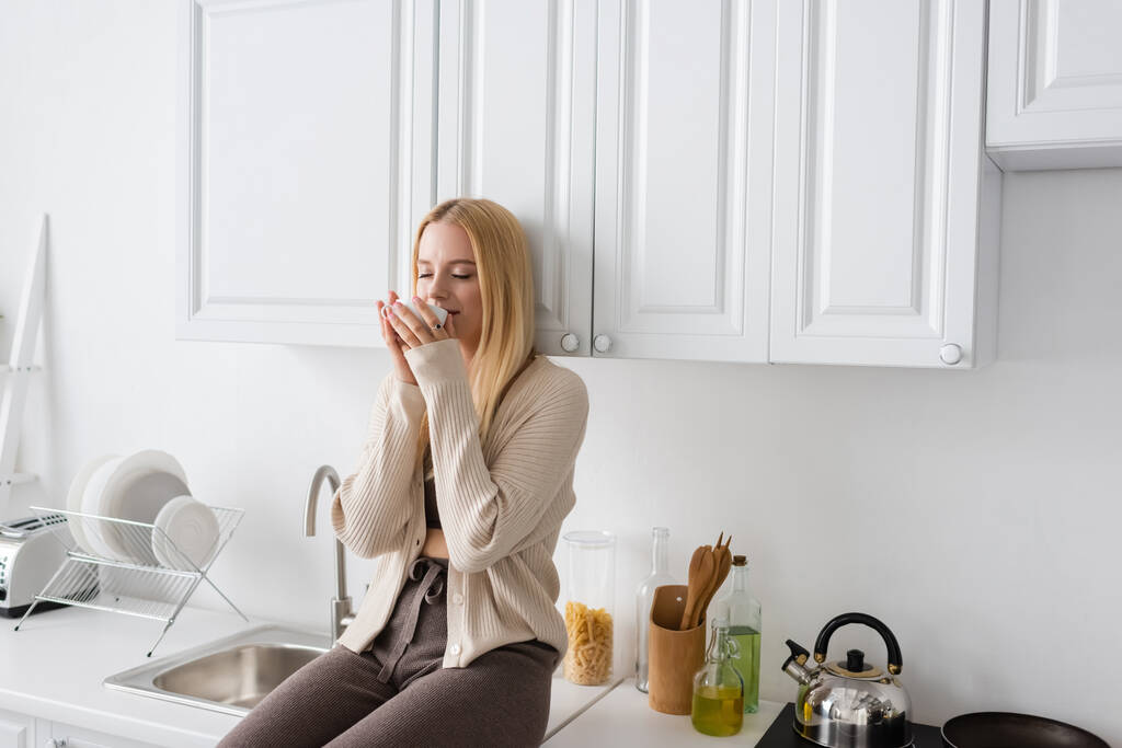 blonde vrouw met gesloten ogen zitten op keuken werkblad en genieten van de smaak van 's ochtends koffie - Foto, afbeelding