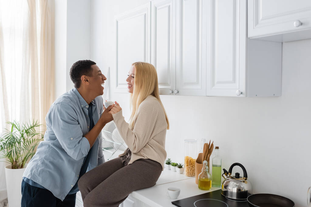 side view of laughing woman sitting on kitchen worktop and holding hands with happy african american boyfriend - Photo, Image