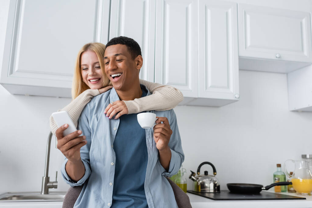 lachender afrikanisch-amerikanischer Mann mit Kaffeetasse und Smartphone neben lächelnder Freundin in der Küche - Foto, Bild
