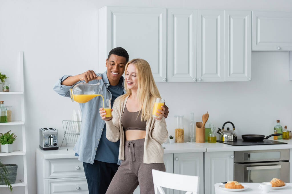 Mutlu Afro-Amerikan adam portakal suyunu güzel kız arkadaşının yanına koyuyor mutfakta bardakları tutuyor. - Fotoğraf, Görsel