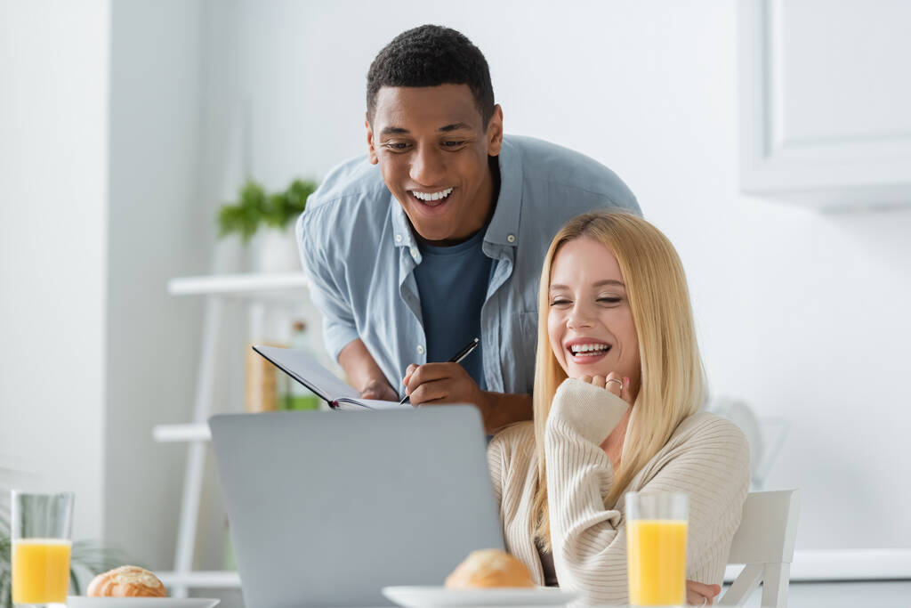 cheerful woman looking at laptop near african american boyfriend with notebook in kitchen - Photo, Image