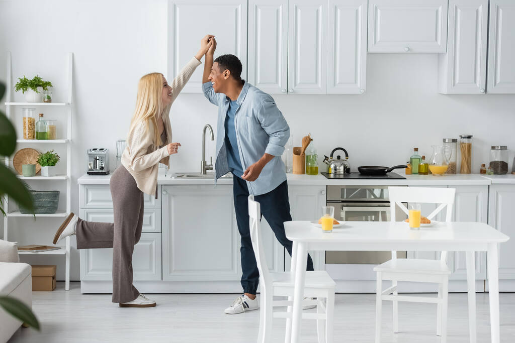 pełna długość wesołej wieloetnicznej pary trzymającej się za ręce i patrzącej na siebie podczas tańca w kuchni - Zdjęcie, obraz