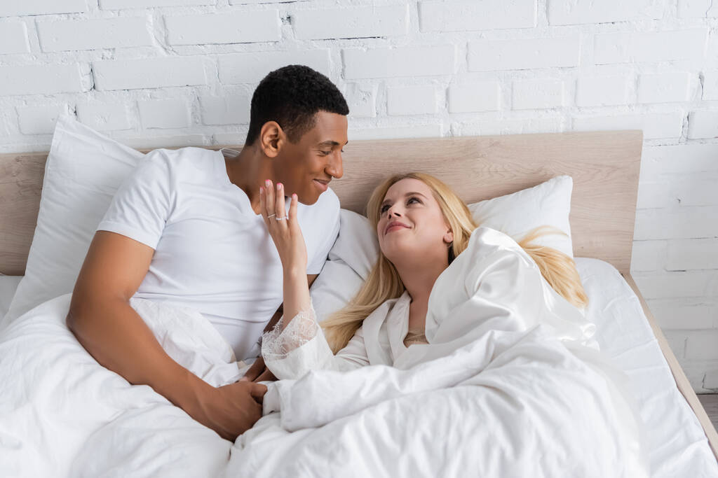 lächelnder afrikanisch-amerikanischer Mann sieht junge und glückliche Freundin an, die morgens sein Gesicht auf dem Bett berührt - Foto, Bild
