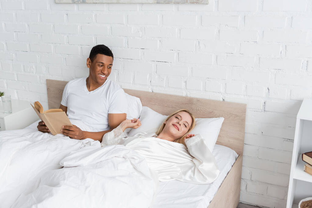 happy african american man with book looking at blonde girlfriend stretching on bed with closed eyes - Photo, Image