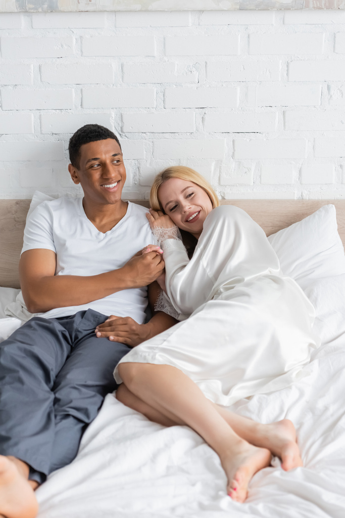 glücklicher afrikanisch-amerikanischer Mann hält die Hand einer blonden Freundin in weißer Robe, die morgens auf dem Bett liegt - Foto, Bild