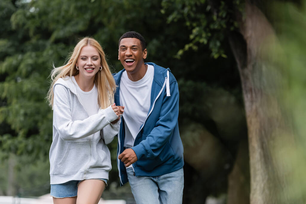 boldog szőke nő és nevető afro-amerikai férfi fogja egymás kezét és sétál a parkban - Fotó, kép