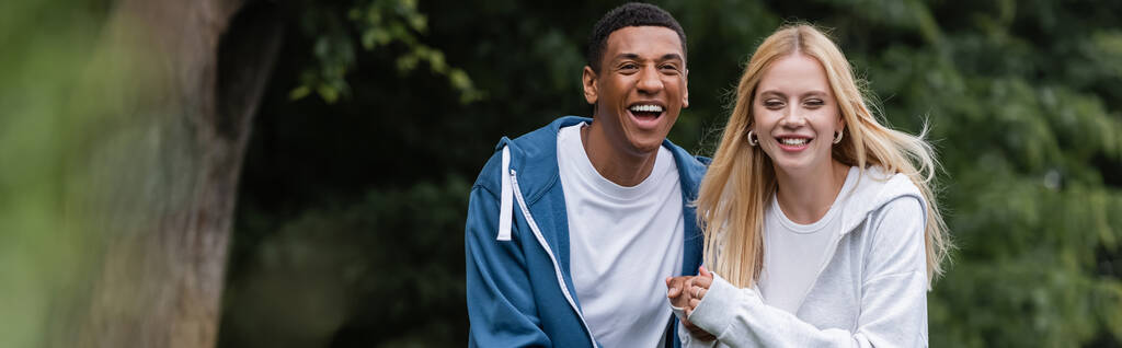radosny Afrykański Amerykanin śmiejący się blisko młoda blondynka dziewczyna w parku, sztandar - Zdjęcie, obraz