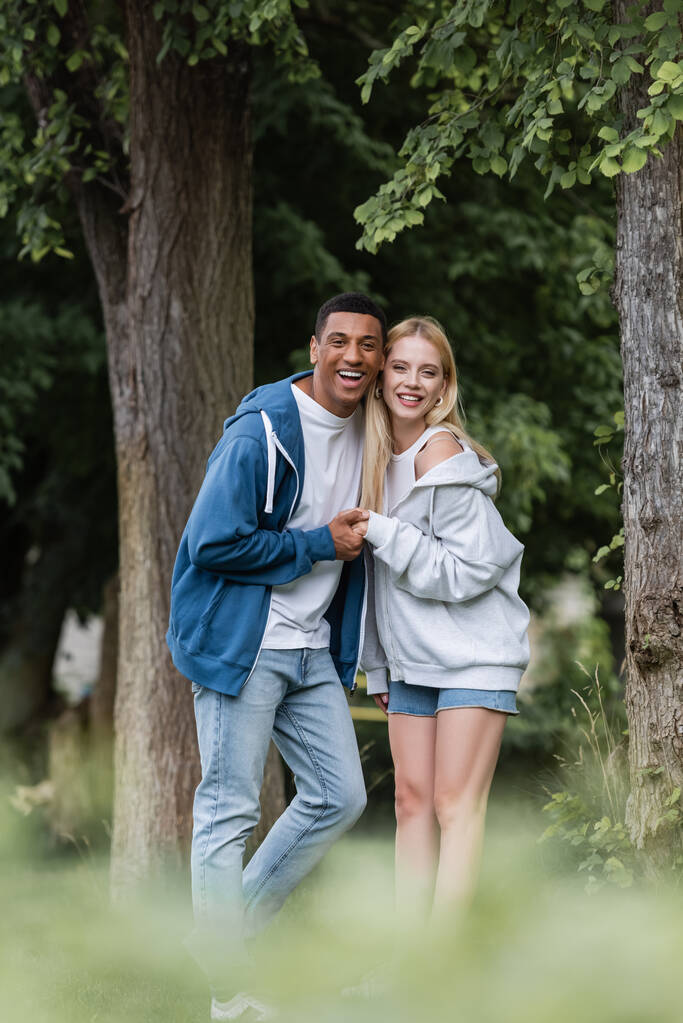 freudiger afrikanisch-amerikanischer Mann und junge blonde Frau in Kapuzenpullis, die sich die Hände halten und im Park in die Kamera schauen - Foto, Bild