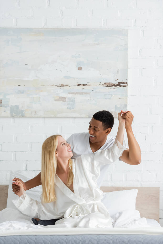 alegre interracial casal segurando as mãos e sorrindo um para o outro enquanto se divertindo no quarto - Foto, Imagem