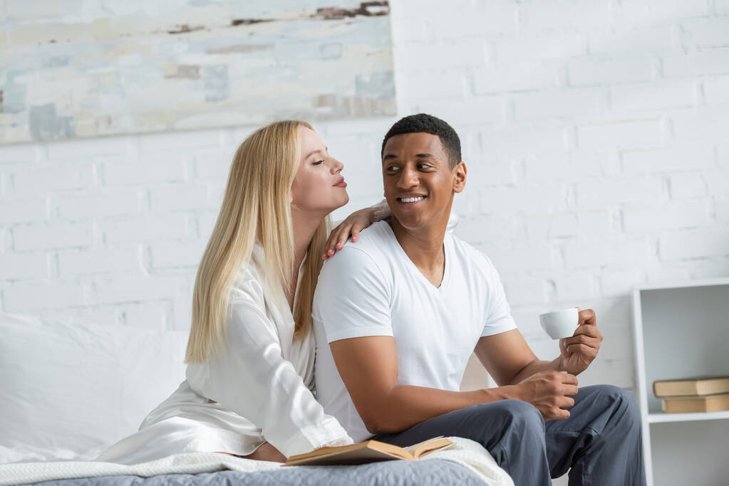 glücklicher afrikanisch-amerikanischer Mann schaut blonde Freundin in weißer Seidenrobe an, während er mit Kaffeetasse im Bett sitzt - Foto, Bild