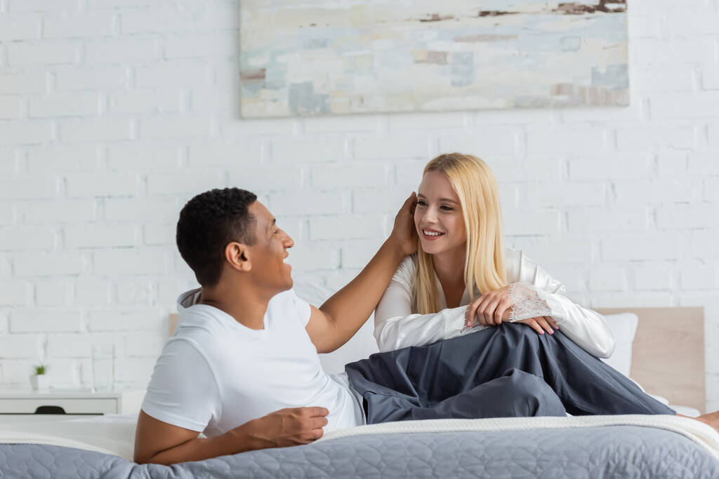 heureux afro-américain toucher les cheveux de jeune copine blonde sur le lit le matin - Photo, image