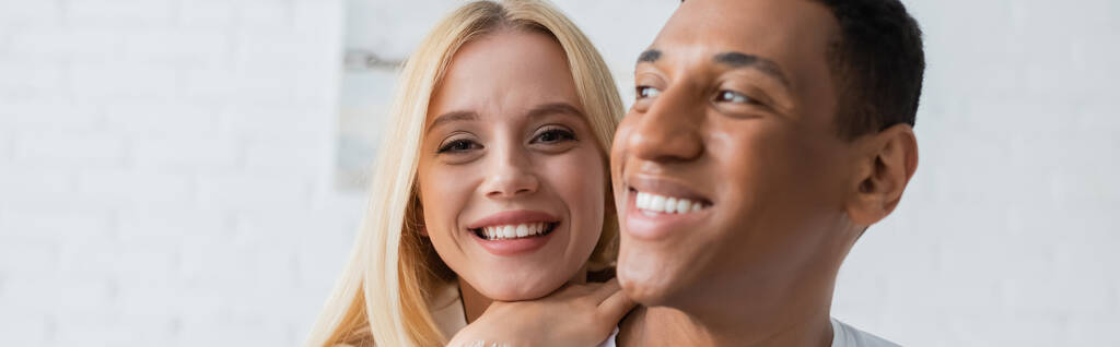 joven y alegre mujer mirando a la cámara cerca de hombre afroamericano sonriendo en primer plano borroso, pancarta - Foto, Imagen