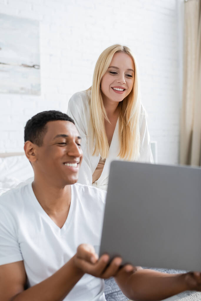 heureux homme afro-américain montrant ordinateur portable à jeune petite amie souriante dans la chambre - Photo, image